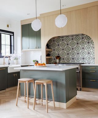 things that make a kitchen look cheap/Scandi style kitchen in sage green and blond wood, tiled backsplash, kitchen island, white pendant lights, bar stools, herringbone floor