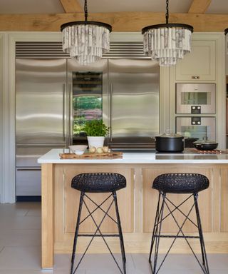 A light wood kitchen with a small peninsula with black barstools and white marble countertops. A huge triple door professional fridge system on the back wall behind the island.
